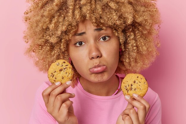 Несчастная молодая женщина с вьющимися волосами не может позволить себе вкусные вкусные угощения, держит два печенья, держится на диете, одержимая выпечкой, одетая в повседневную футболку, изолированную на розовом фоне