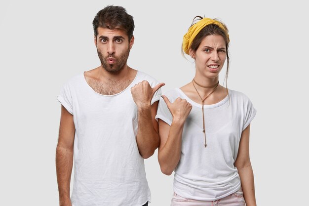 Несчастная молодая женщина с европейской внешностью, показывает большим пальцем на удивленного парня, выражает неприязнь и удивление, носит повседневные футболки, изолирована на белой стене