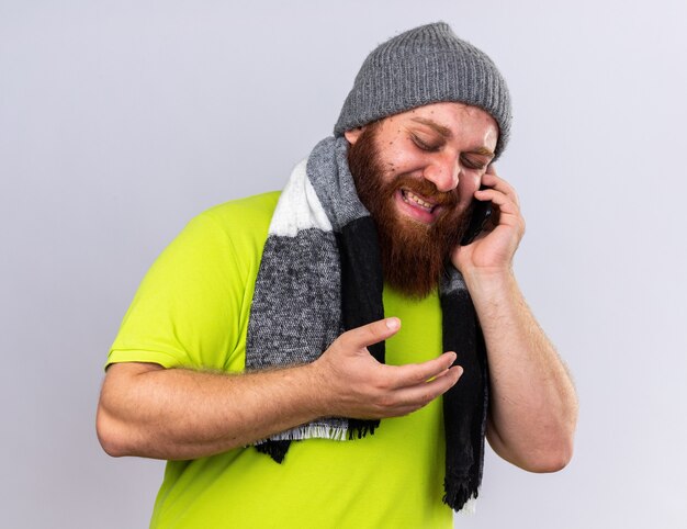 Unhealthy bearded man in hat and with warm scarf around neck feeling sick suffering from flu talking on mobile phone looking disappointed standing over white wall