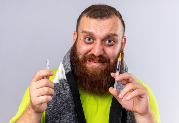 Foto gratuita uomo barbuto malsano con sciarpa calda intorno al collo che si sente male soffre di influenza tenendo la siringa e l'ampolla che sembra preoccupato e spaventato