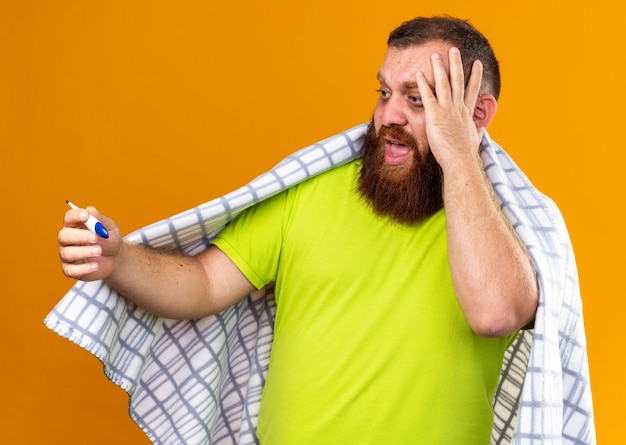 Unhealthy bearded man wrapped in blanket feeling sick suffering from cold checking temperature using thermometer being worried and scared