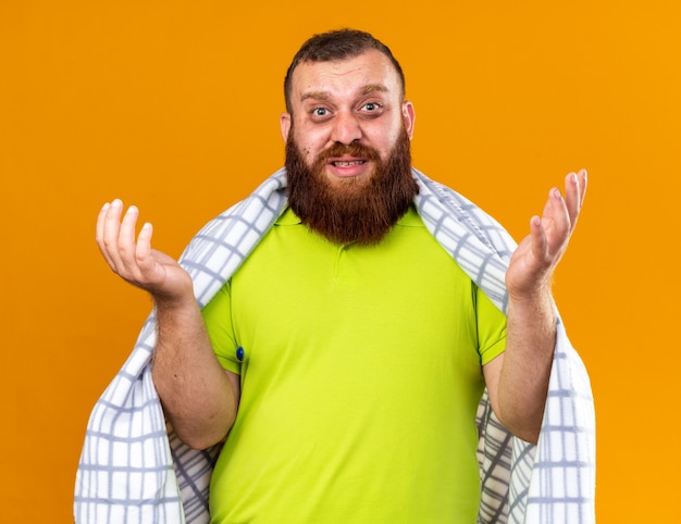 Free photo unhealthy bearded man wrapped in blanket feeling sick suffering from cold checking temperature using thermometer looking confused with arms raised