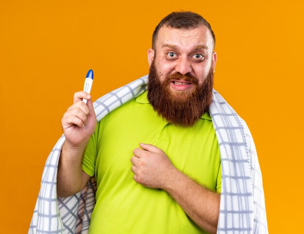 Unhealthy bearded man wrapped in blanket feeling sick suffering from cold checking temperature using thermometer looking worried and confused standing over orange wall