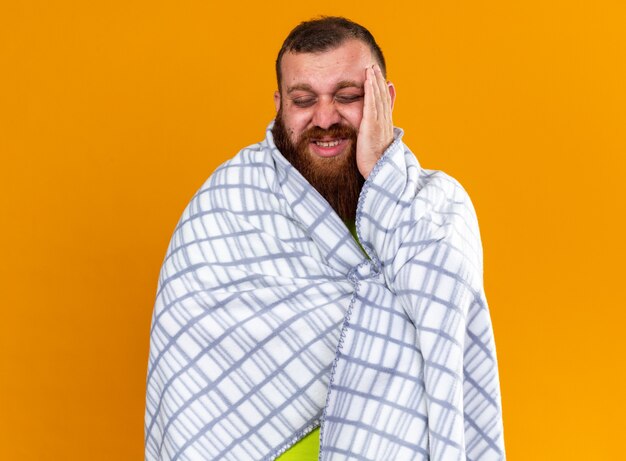 Нездоровый бородатый мужчина, завернутый в одеяло, чувствует себя больным, страдает от простуды и сильной головной боли
