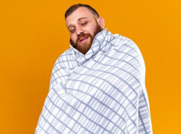 Нездоровый бородатый мужчина, завернутый в одеяло, чувствует себя больным, страдает от холода, с закрытыми глазами стоит над оранжевой стеной