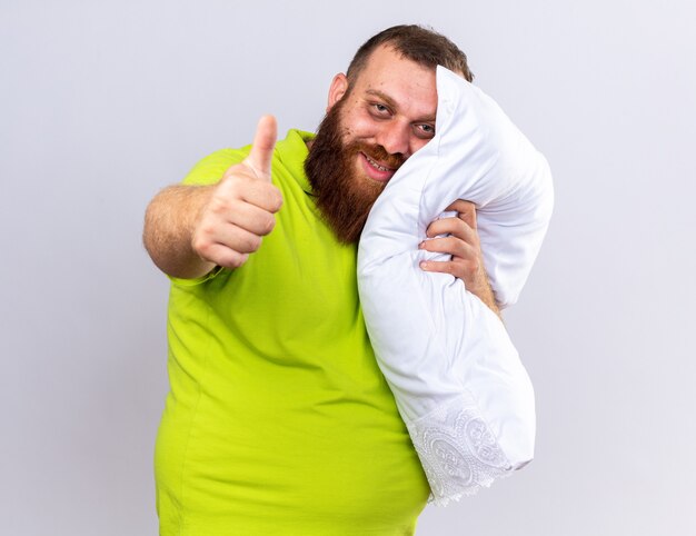 Unhealthy bearded man in yellow polo shirt feeling better holding pillow  smiling showing thumbs up standing over white wall