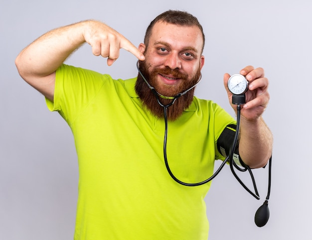 Free photo unhealthy bearded man in yellow polo shirt feeling better measuring blood pressure using tonometer  pointing with index finger at it smiling standing over white wall