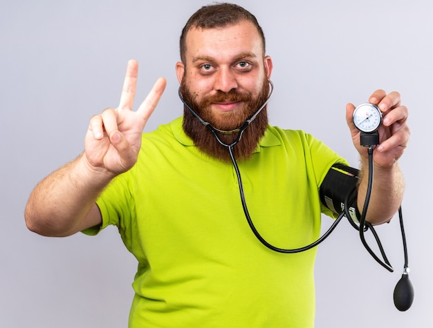 Нездоровый бородатый мужчина в желтой рубашке-поло чувствует себя лучше, измеряя артериальное давление с помощью тонометра, улыбаясь, показывая знак v, стоящий над белой стеной