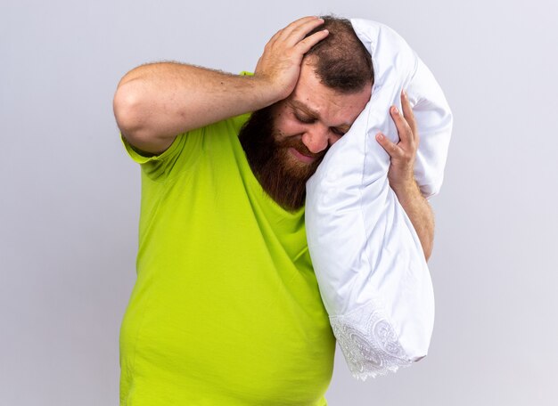 Unhealthy bearded man in yellow polo shirt feeling sick holding pillow suffering from strong headache standing over white wall