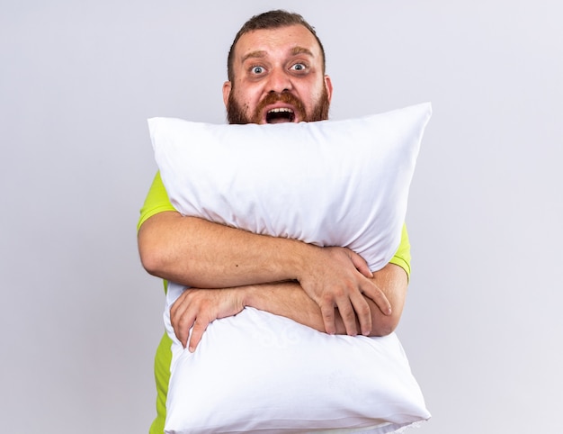 Unhealthy bearded man in yellow polo shirt feeling sick hugging pillow  shouting with annoyed expression standing over white wall