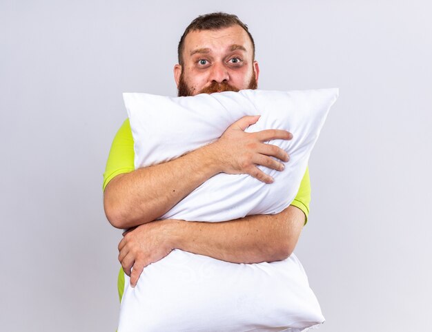 Unhealthy bearded man in yellow polo shirt feeling sick hugging pillow  with sad expression standing over white wall