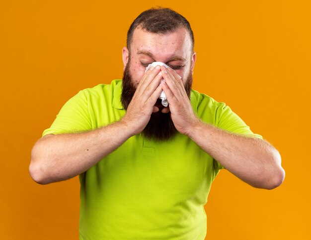 Unhealthy bearded man in yellow polo shirt feeling terrible suffering from cold blowing running nose sneezing in tissue standing over orange wall