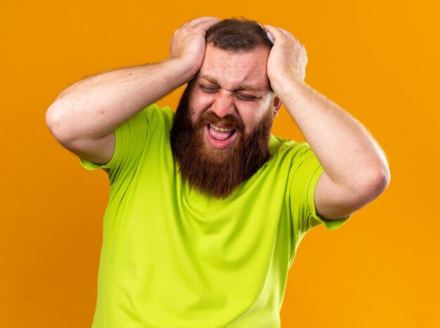 Unhealthy bearded man in yellow polo shirt feeling terrible suffering from cold and strong headache touching his head