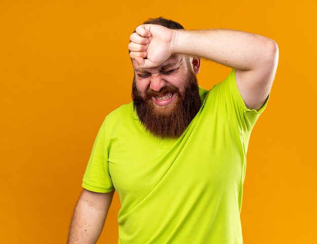 노란색 폴로 셔츠를 입은 건강에 해로운 수염을 가진 남자는 심한 두통을 느끼는 고통으로 고통받는 이마를 만져 추위로 고통받습니다.