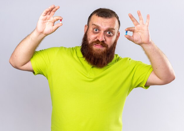 Unhealthy bearded man in yellow polo shirt holding different pills feeling sick suffering from flu   confused