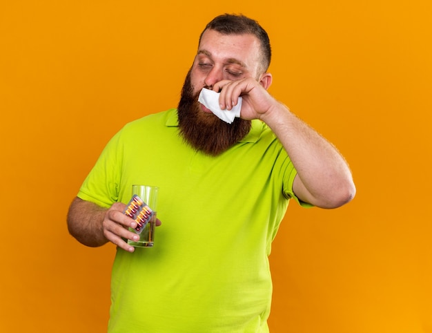 Нездоровый бородатый мужчина в желтой рубашке-поло со стаканом воды и таблетками, чувствуя ужасное сморкание, насморк, чихание тканями, страдающий от холода, стоя над оранжевой стеной
