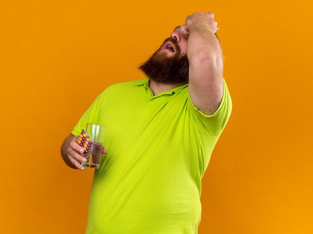 Нездоровый бородатый мужчина в желтой рубашке поло, держащий стакан воды и таблетки, чувствует себя ужасно, страдая от простуды и сильной головной боли, стоя над оранжевой стеной
