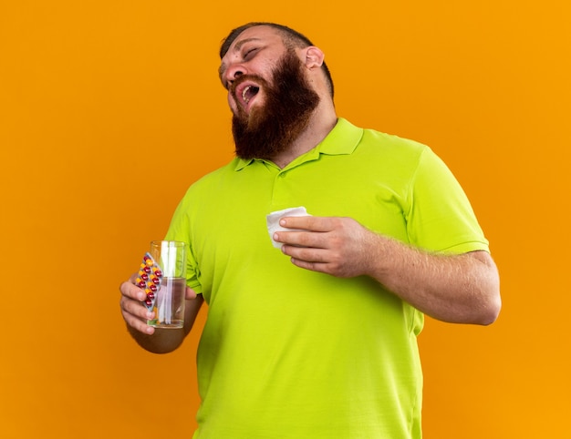 Unhealthy bearded man in yellow polo shirt holding glass of water and pills feeling terrible suffering from cold going to sneeze standing over orange wall