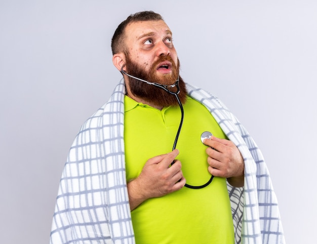 Foto gratuita barbuto malsano avvolto in una coperta che si sente male soffre di freddo ascoltando il suo battito cardiaco usando uno stetoscopio che sembra preoccupato