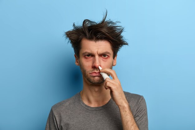 Unhealthy man treats nose with spray, suffers from allergic rhinitis, has watery red eyes, first symptoms of virus, no addictions to medicals, isolated over blue wall. Sinusitis treatment