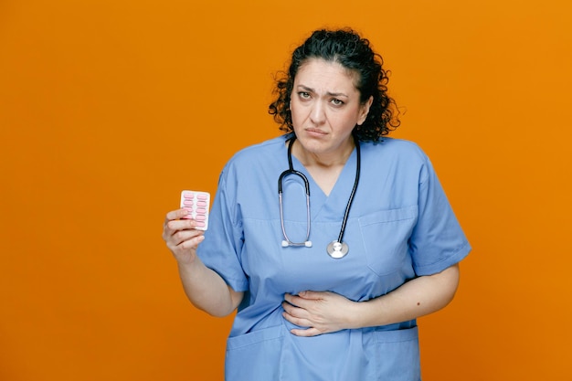 Нездоровая женщина-врач средних лет в униформе и со стетоскопом на шее показывает упаковку капсул, смотрит в камеру, держа руку на животе, изолированную на оранжевом фоне