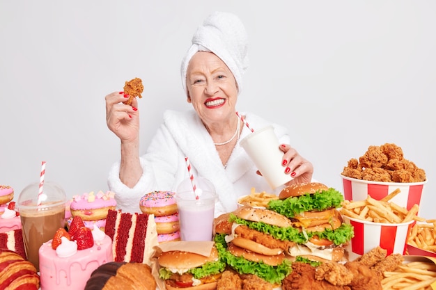 Free photo unhealthy overeating lifestyle. pleased old lady smiles positively drinks soda eats fast food