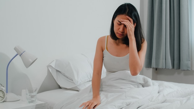 Unhealthy sick Indian female suffers from insomnia. Asian young woman taking painkiller medicine to relieve headache pain and drink glass of water sitting on bed in her bedroom at home in morning.