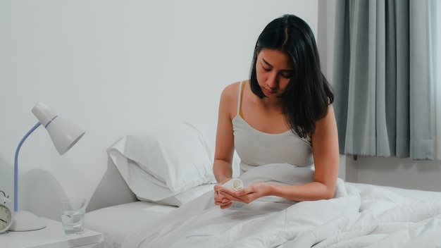 Unhealthy sick Indian female suffers from insomnia. Asian young woman taking painkiller medicine to relieve headache pain and drink glass of water sitting on bed in her bedroom at home in morning.