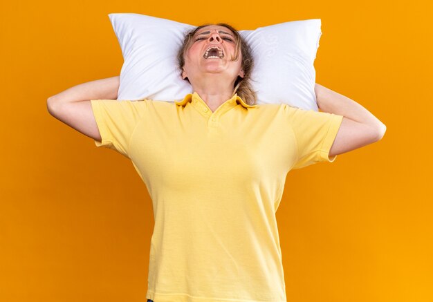 Unhealthy woman suffering from flu and cold with pillow behind her head over orange wall