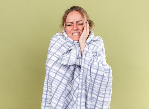 Donna malsana avvolta in una coperta che si sente terribilmente affetta da influenza e raffreddore con forte mal di testa in piedi sul muro verde