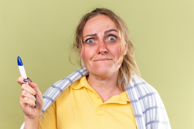 Foto gratuita donna malsana avvolta in una coperta che si sente terribilmente sofferente di influenza e termometro che tiene freddo e sembra preoccupata in piedi sul muro verde