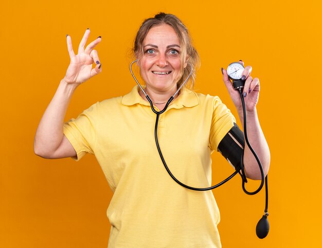 Нездоровая женщина в желтой рубашке чувствует себя лучше, измеряя свое кровяное давление с помощью тонометра, улыбаясь, показывая знак ОК, стоящий над оранжевой стеной