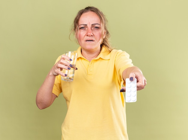 노란색 셔츠에 건강에 해로운 여자는 독감으로 고통받는 물과 약의 잔을 들고 끔찍한 느낌