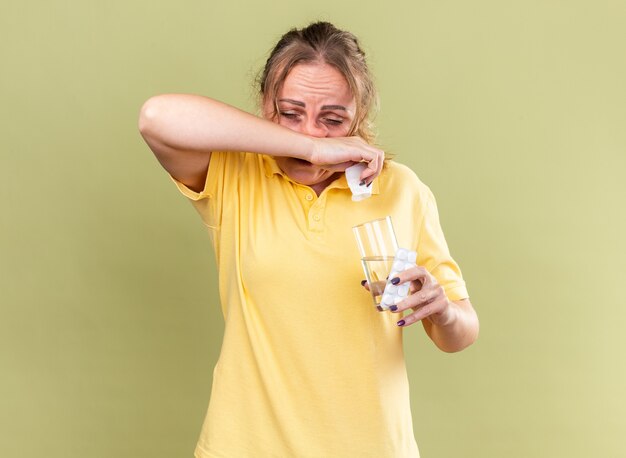 Unhealthy woman in yellow shirt feeling terrible holding glass of water and pills wiping running nose suffering from flu standing over green wall