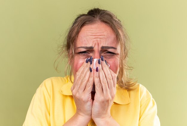 Нездоровая женщина в желтой рубашке чувствует себя ужасно, страдая от гриппа и холодного сморкания, насморка, чихания тканью