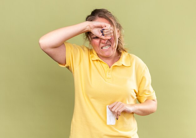 Unhealthy woman in yellow shirt feeling terrible touching her forehead suffering from strong headache because of blocked nose having flu standing over green wall