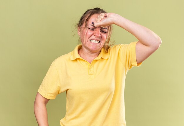 Unhealthy woman in yellow shirt feeling terrible touching her forehead suffering from strong headache having flu standing over green wall