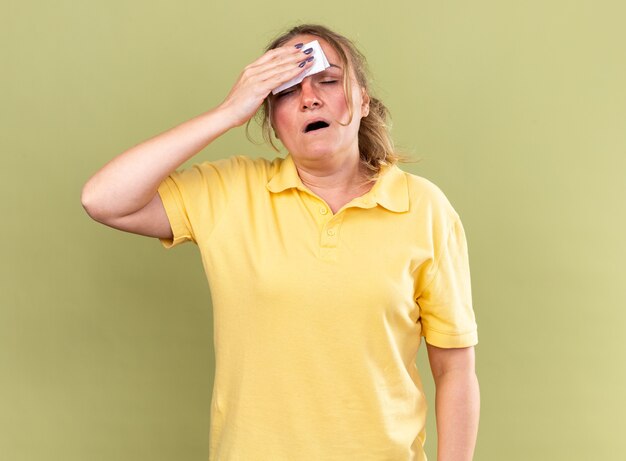 Нездоровая женщина в желтой рубашке чувствует себя ужасно, вытирая лоб салфеткой, болеет гриппом и лихорадкой