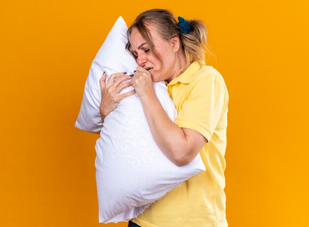 Unhealthy woman in yellow shirt feeling unwell suffering from flu and cold hugging pillow coughing standing over orange wall