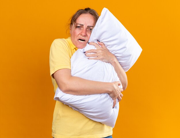 Unhealthy woman in yellow shirt feeling unwell suffering from flu and cold hugging pillow standing over orange wall