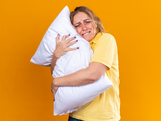 黄色いシャツを着た不健康な女性がインフルエンザにかかり、オレンジ色の壁の上に立つ熱に苦しんで冷たい抱き枕を抱きしめる