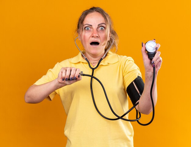黄色いシャツを着た不健康な女性がインフルエンザや風邪にかかり、オレンジ色の壁の上に立って心配している眼圧計を使って血圧を測定している