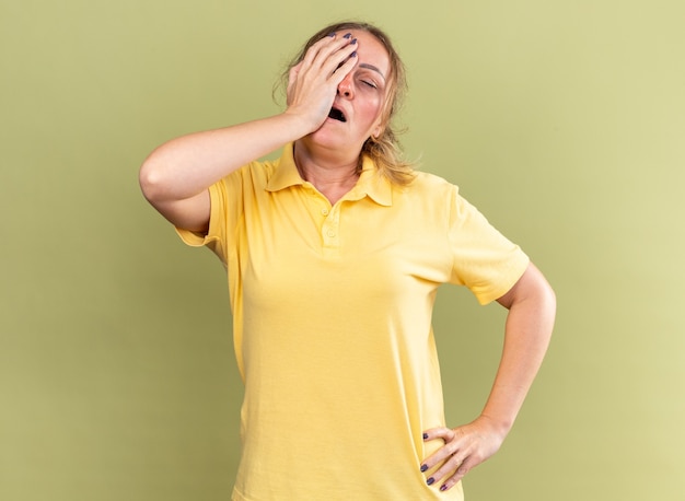 Нездоровая женщина в желтой рубашке чувствует себя нездоровой, касаясь лба, когда у нее кружится голова от гриппа, стоящего у зеленой стены