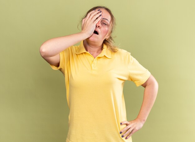 Unhealthy woman in yellow shirt feeling unwell touching her forehead while feeling dizzy having flu standing over green wall