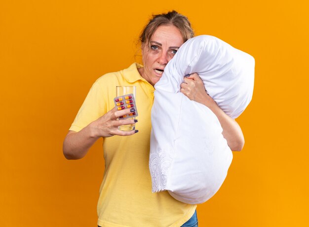 Unhealthy woman in yellow shirt suffering from flu and cold feeling unwell hugging pillow holding pills and glass of water standing over orange wall