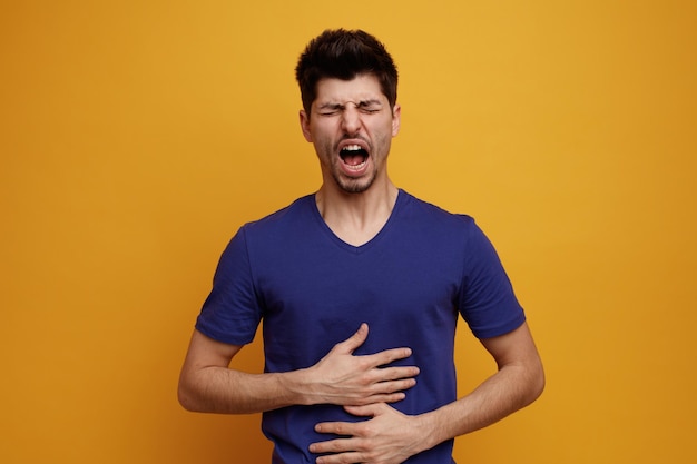 Unhealthy young handsome man keeping hands on belly screaming with closed eyes on yellow background