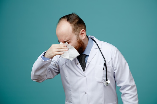 Бесплатное фото Нездоровый молодой врач-мужчина в медицинском халате и со стетоскопом на шее вытирает нос салфеткой с закрытыми глазами на синем фоне