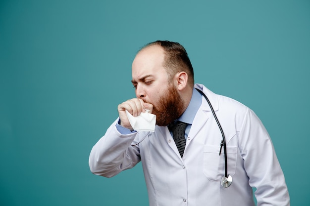 Unhealthy young male doctor wearing medical coat and stethoscope around his neck holding napkin coughing with closed eyes isolated on blue background