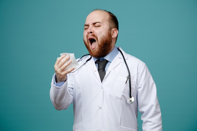 Unhealthy young male doctor wearing medical coat and stethoscope around his neck holding napkin sneezing with closed eyes isolated on blue background