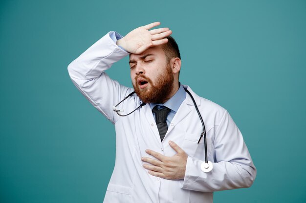 Unhealthy young male doctor wearing medical coat and stethoscope around his neck keeping hand on chest and on head with closed eyes isolated on blue background